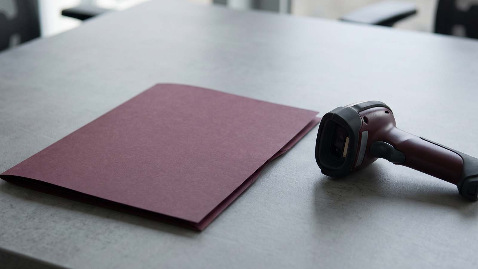 A closed burgundy folder lying next to a handheld barcode scanner on a grey desk.