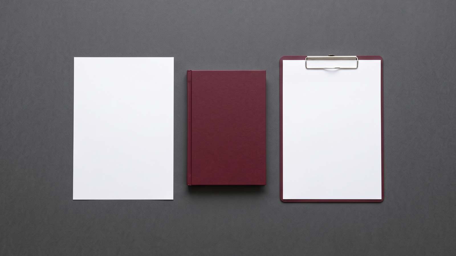 A blank sheet of paper, a closed burgundy notebook and a blank clipboard laid out side by side on a dark grey surface.